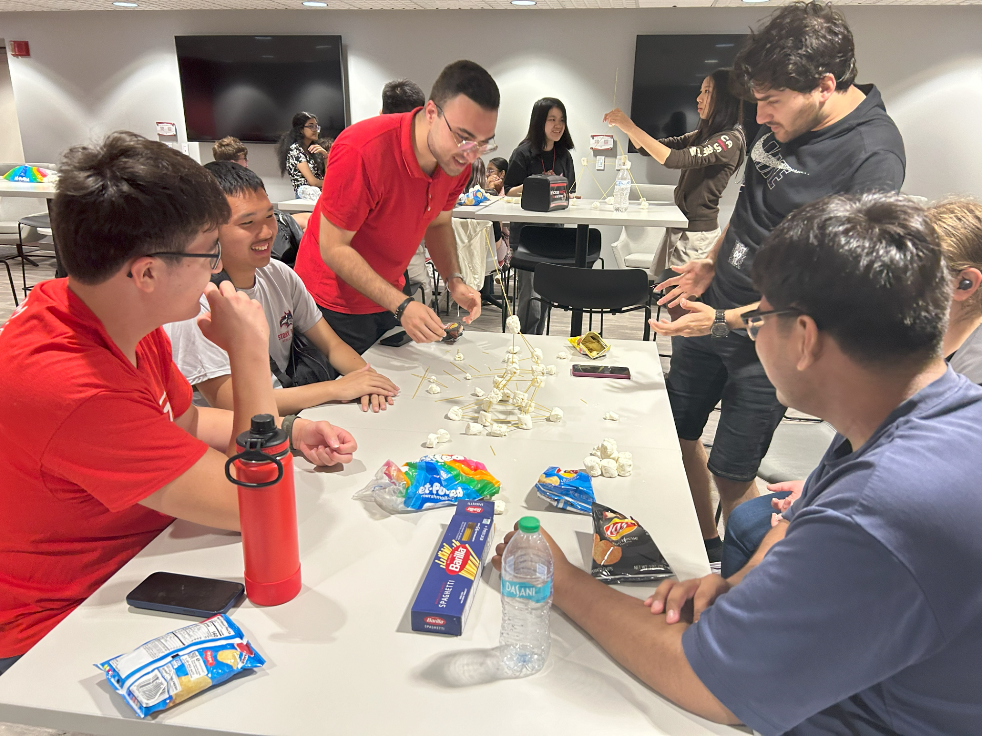 Students sitting at a desk with marshmallows and pasta at an event.