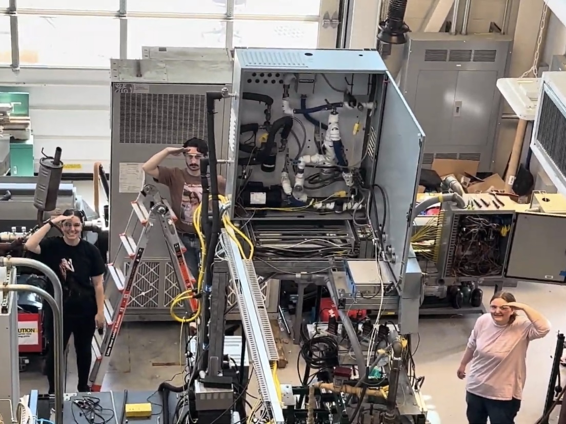 A group of students saluting while working in a lab with machines.