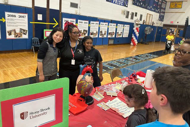Educators and a child at a school fair, standing by a table with a heart model and Stony Brook University sign.