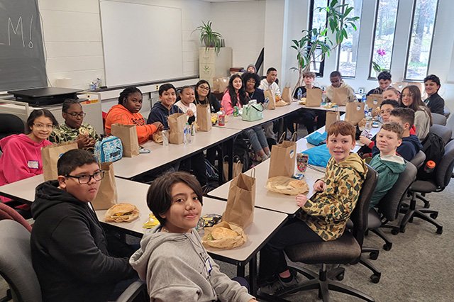A diverse group of students are sitting around a U-shaped table in a classroom, smiling and enjoying packed lunches.