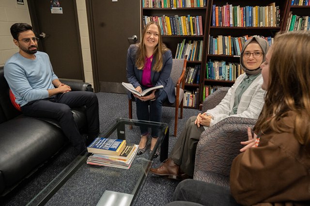 Professor Elizabeth Hewitt is seated in a library and surrounded by four students, and she is smiling with a book in her hand