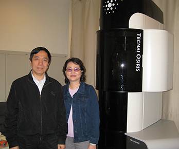 Two individuals stand smiling beside a black and white Tecnai Osiris machine in a room.
