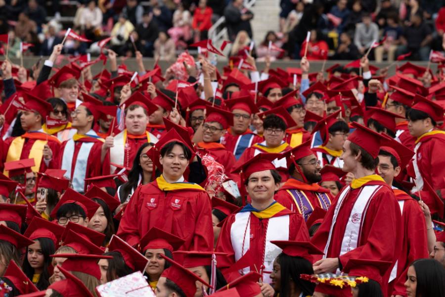 A large group of graduates in red caps and gowns celebrate, many smiling and holding small flags.
