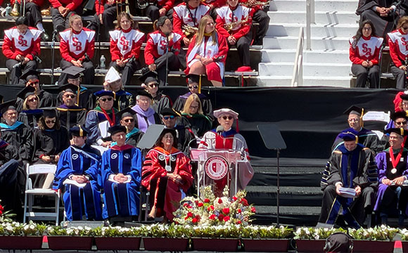 A graduation ceremony with a speaker at a podium and faculty seated in academic regalia.