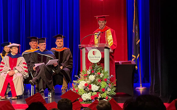 A speaker in red academic regalia at a podium with seated faculty members at a graduation ceremony.