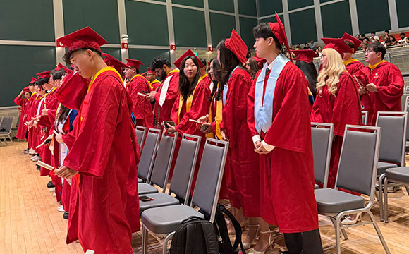Graduates in red gowns and caps stand in an auditorium with green walls and wooden floors.