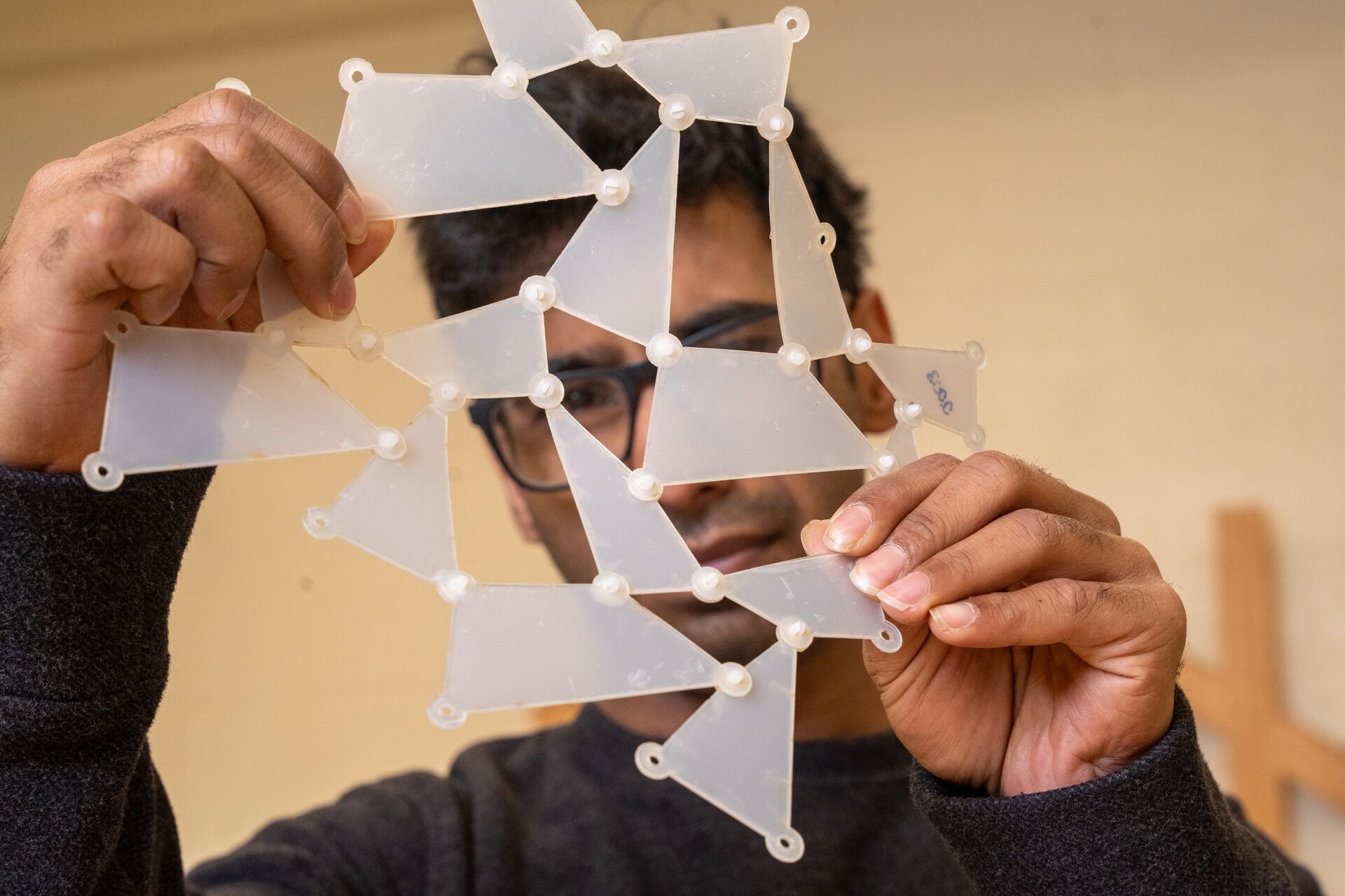 A man in glasses holds a translucent, flexible geometric structure made of triangular panels connected by small nodes, conveying curiosity and creativity.