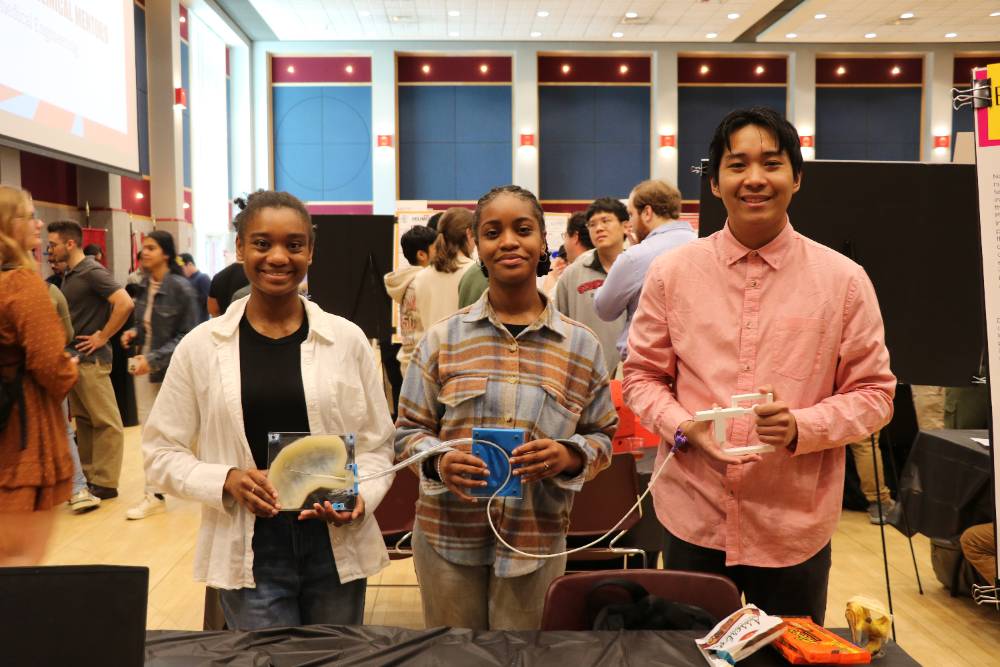 Three young adults stand smiling in a busy exhibition hall, each holding a unique crafted item, suggesting a creative or educational event.