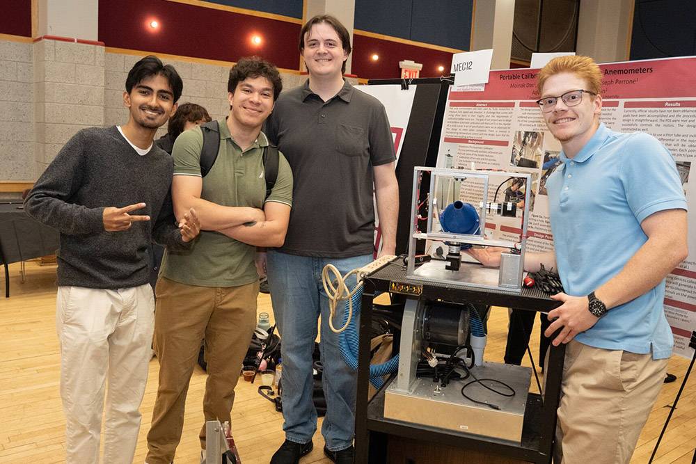Four people stand smiling beside a mechanical project display in a room with informational posters.