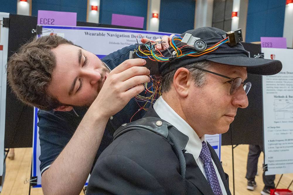 A man adjusts a wearable navigation device on another man's cap at a tech expo. Colorful wires and sensors are visible.