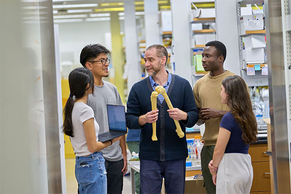 A group of five individuals in a lab setting discusses a scientific topic, with a man holding a model of human bones at the center.