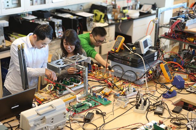 Electric Energy Lab Three individuals work intently on electronic equipment in a cluttered lab, surrounded by wires, circuits, and measurement tools, conveying focus and collaboration.