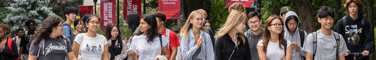 A diverse group of students walking on a campus with trees and banners in the background