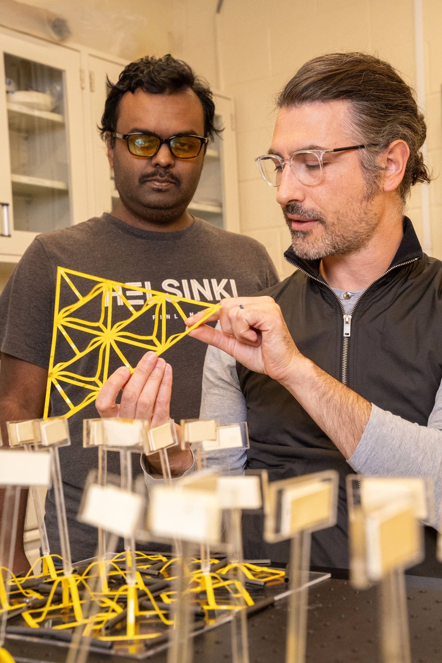 Two researchers in a lab setting are examining a bright yellow, 3D-printed geometric lattice structure. The person in the foreground holds the intricate triangular-patterned frame, while the other researcher looks on. In the lower foreground, several similar yellow structures are mounted on small stands, each featuring a small rectangular sample plate, likely for testing material properties.