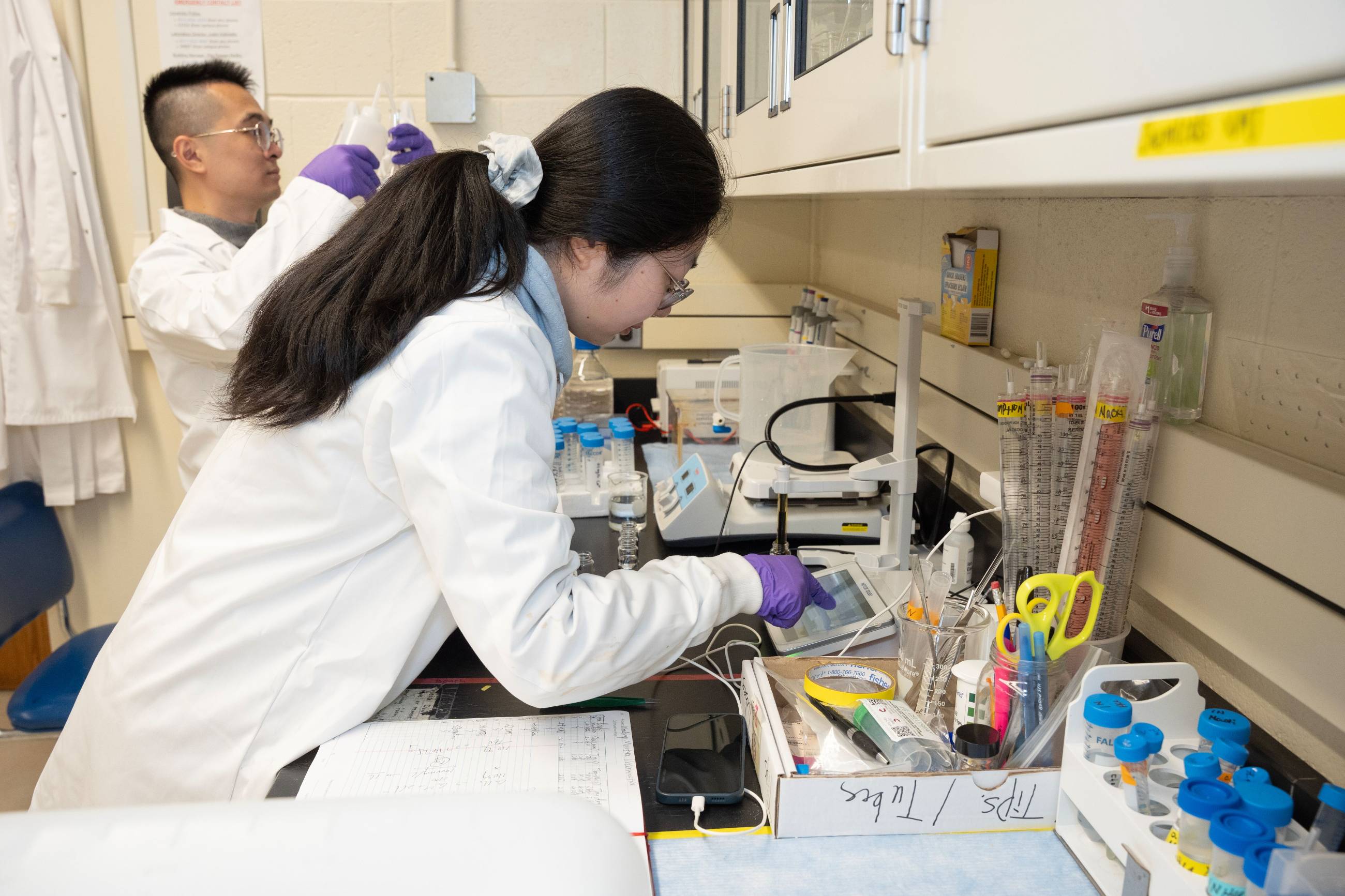 Two researchers wearing white lab coats and purple nitrile gloves work in a busy laboratory. In the foreground, a student leans over a workbench, carefully adjusting settings on a digital device while consulting a notebook. In the background, a second student is seen handling laboratory glassware. The lab is filled with scientific equipment, including test tubes, pipettes, and graduated cylinders.