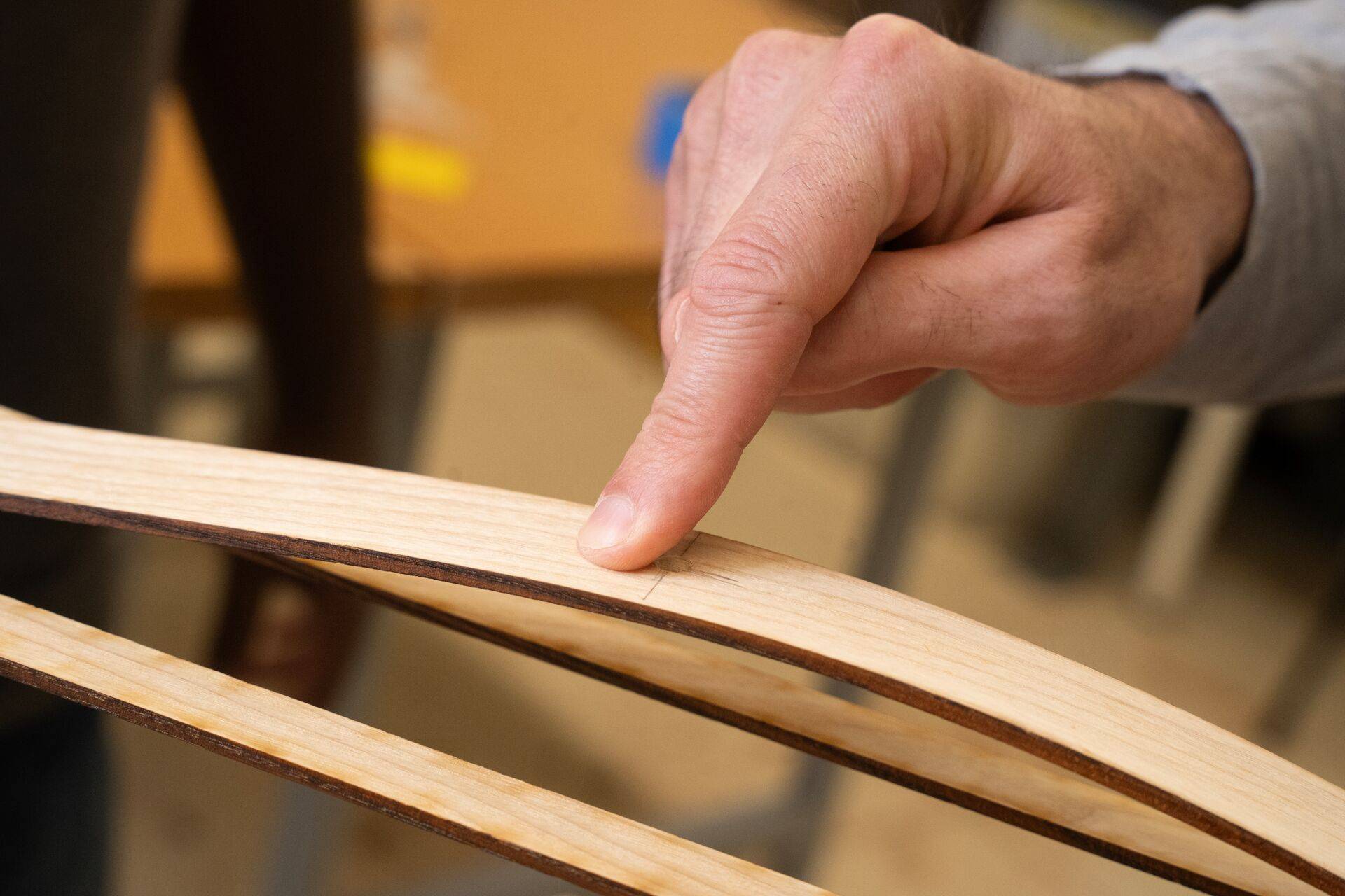 A close-up shot of a person's finger pointing to a small, precise mark on a thin, curved strip of light-colored wood. The wood appears to be part of a larger structure, in an engineering lab setting.