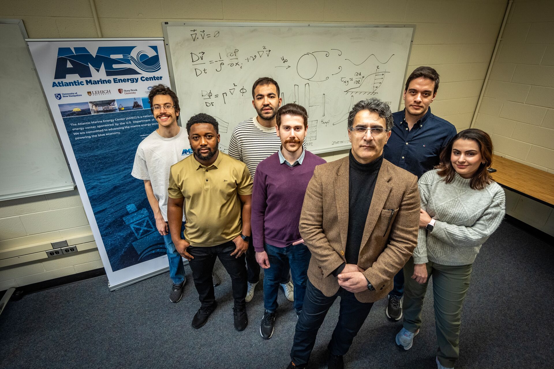 A group portrait of a diverse research team standing in front of a whiteboard filled with complex fluid dynamics equations and diagrams. To their left is a banner for the Atlantic Marine Energy Center (AMEC), which lists partner institutions including Stony Brook University. The lead researcher, in a brown blazer and black turtleneck, stands at the front, surrounded by five students or junior researchers.
