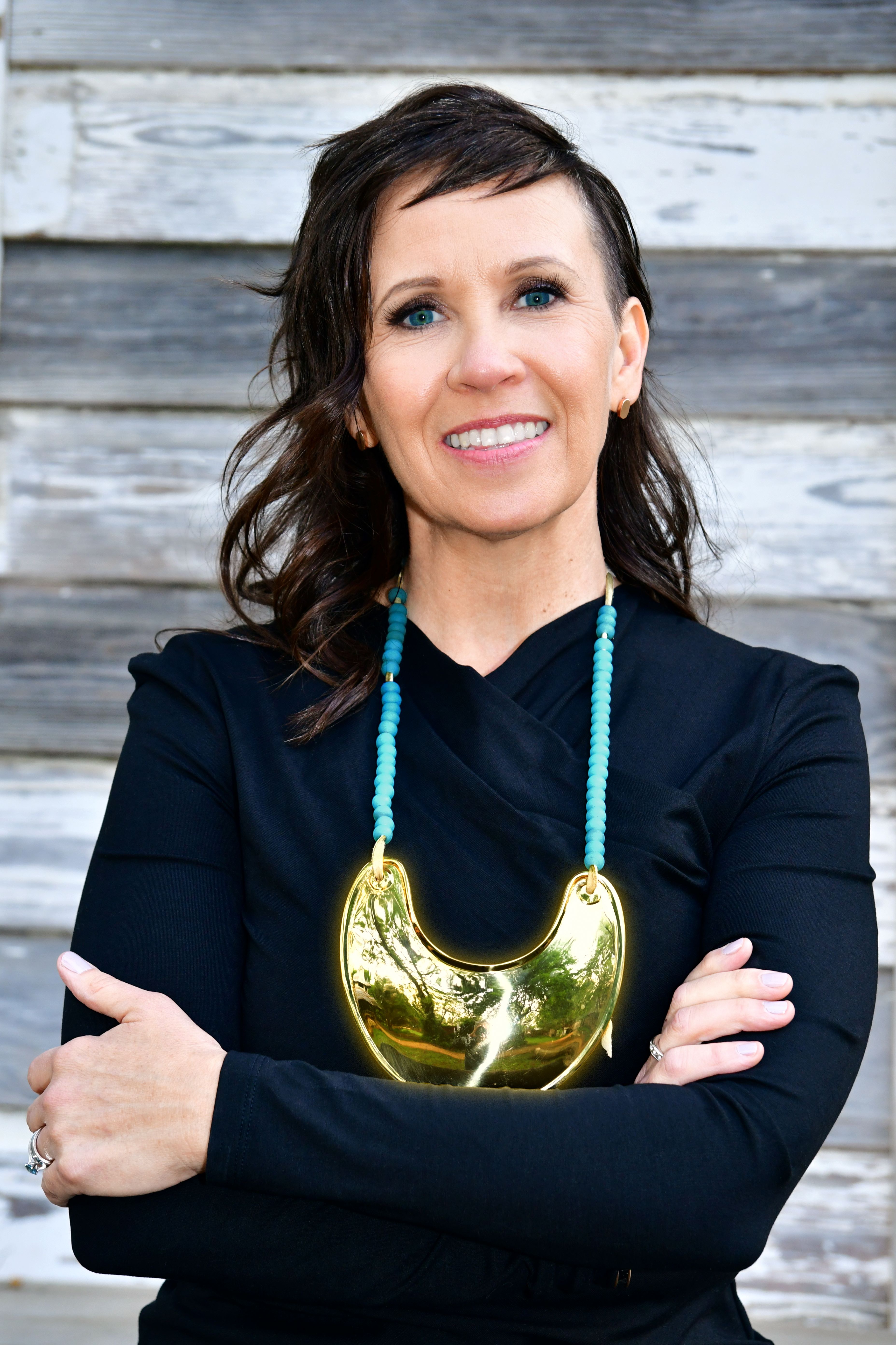 Person wearing a black top and a large gold necklace, smiling in front of a wooden plank background.