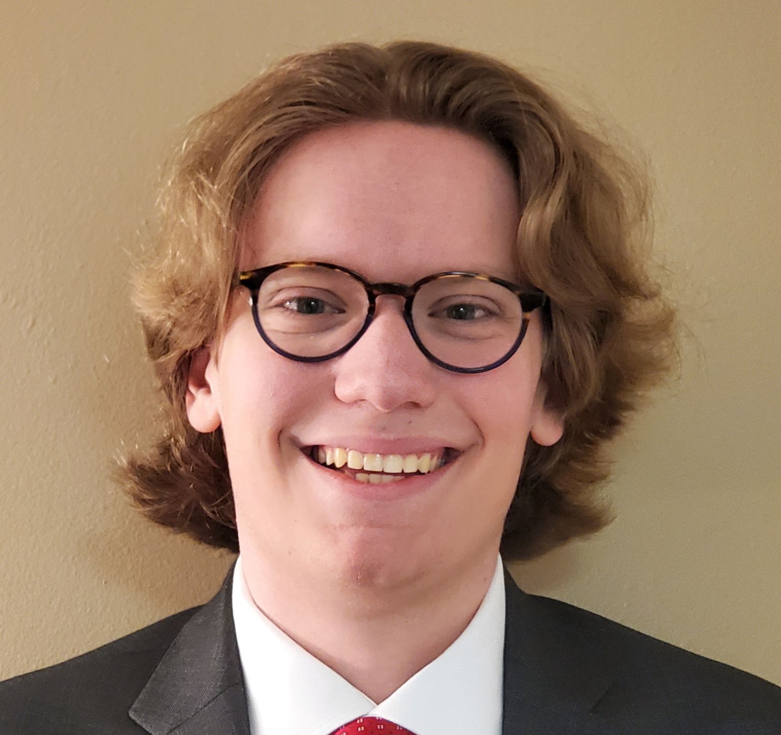 Matthew Jacob Portrait of a smiling person with shoulder-length curly hair, wearing glasses, a red tie, and a dark jacket.
