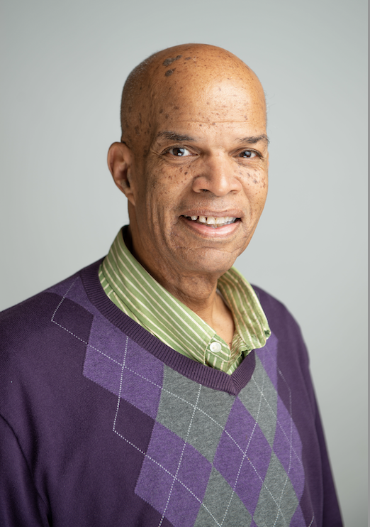 Portrait of a smiling individual wearing a purple and gray argyle sweater over a green striped shirt, against a light gray background.