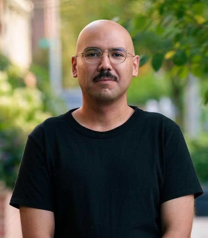 Portrait of an individual with a shaved head wearing a black T-shirt, standing outdoors with greenery in the background.