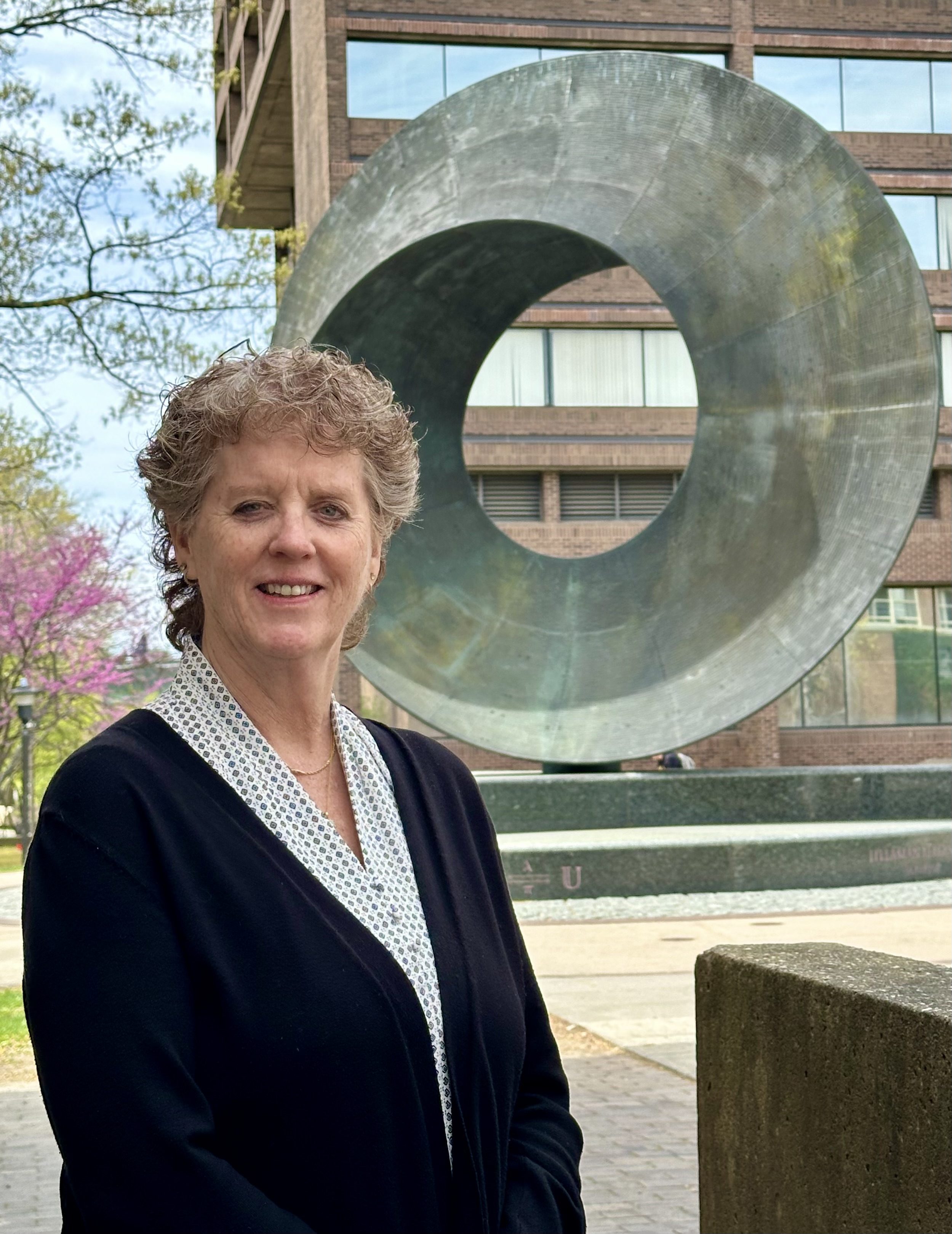 Portrait of an individual smiling, standing outdoors in front of a sculpture of a torus