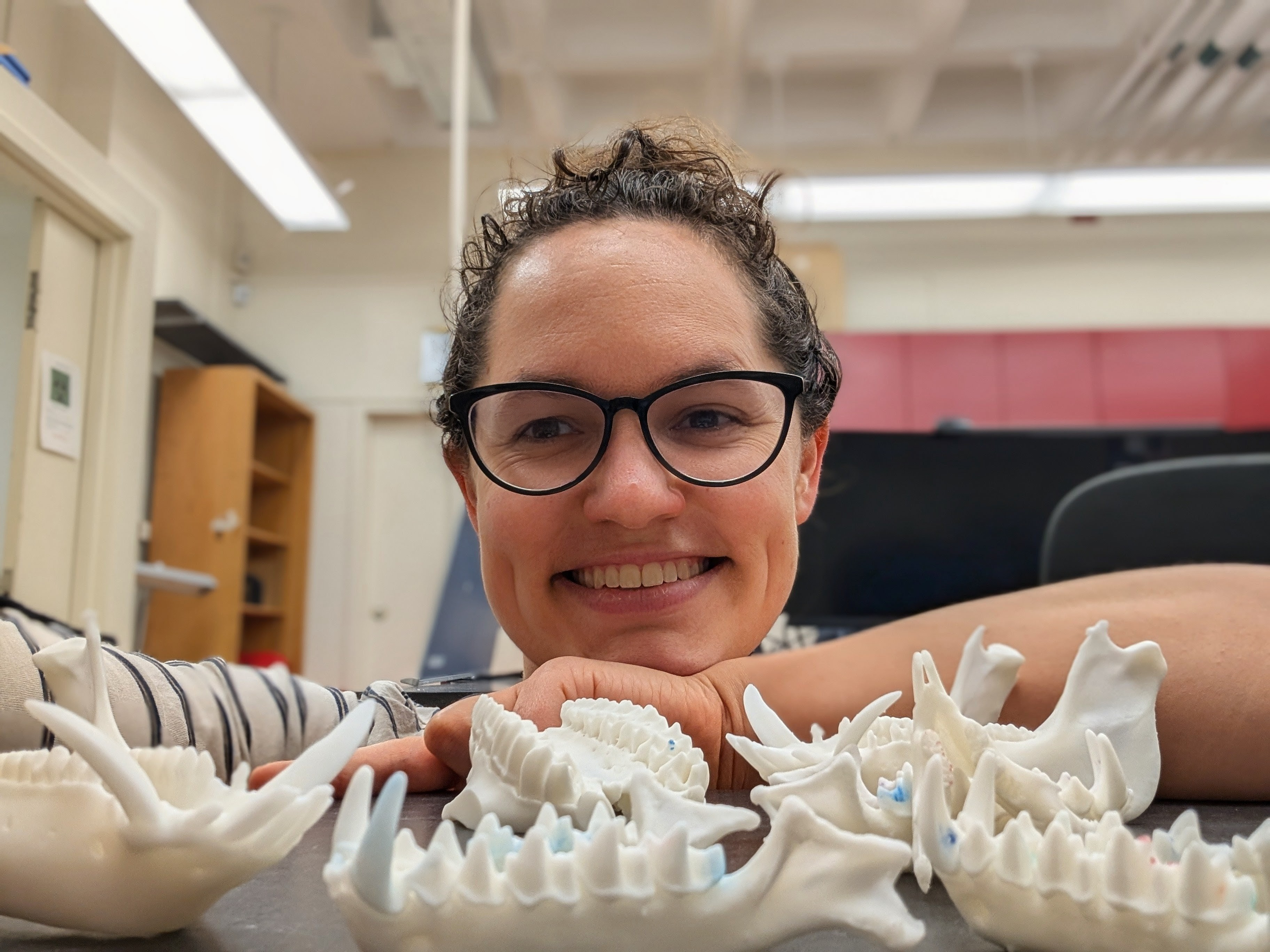 A smiling person wearing eyeglasses stands in front of fossils that lay on a table, with her hands under her chin