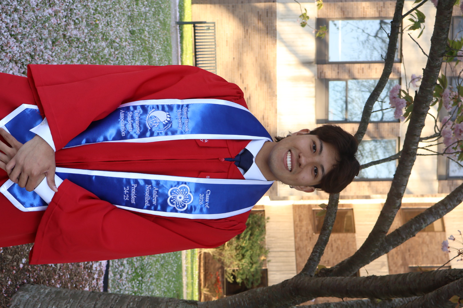 A graduate in a red gown and blue stole smiles under a blooming tree. The background shows a brick building and lawn