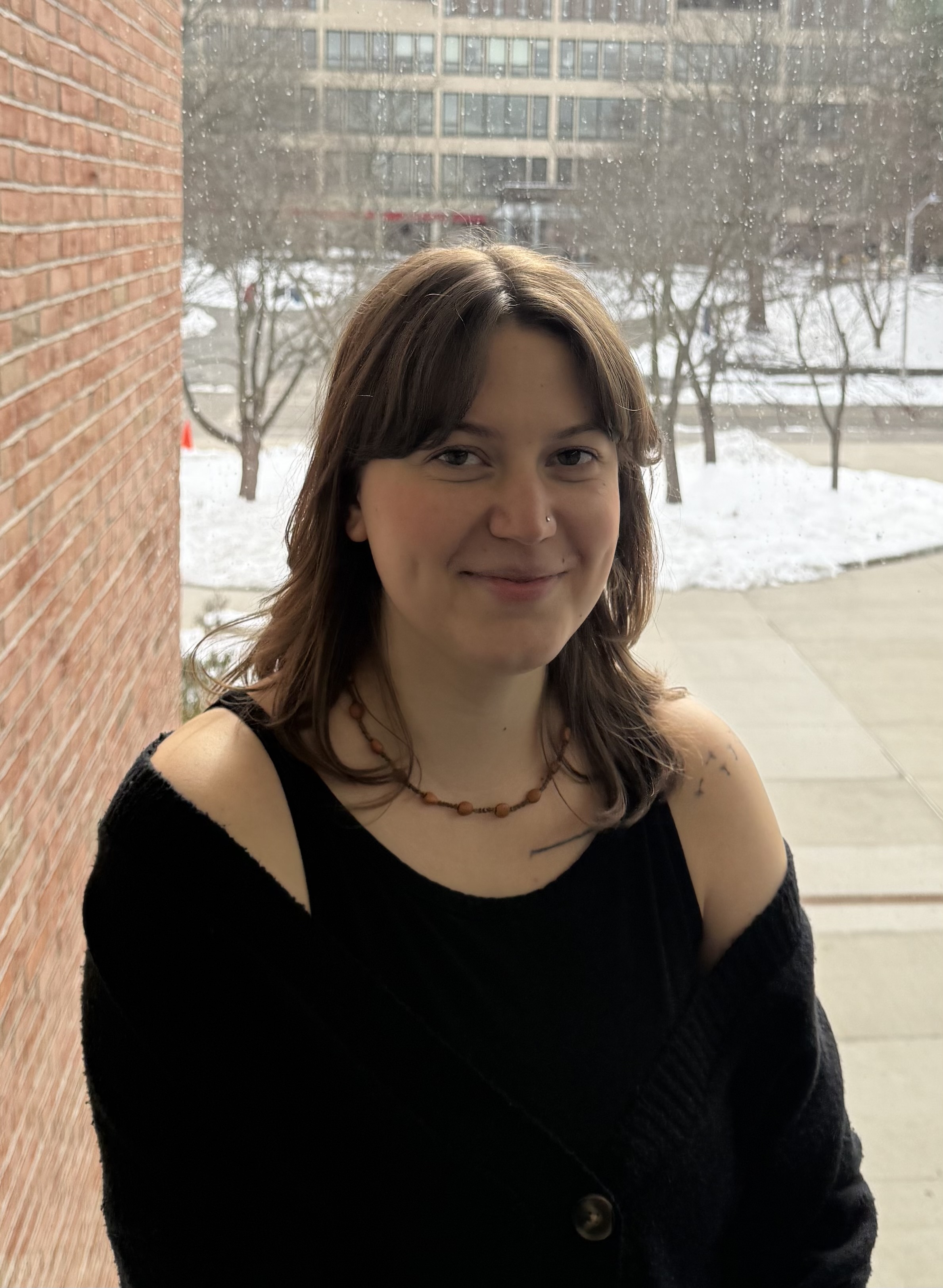 A person with shoulder-length hair smiles softly, standing against a snowy outdoor backdrop. 