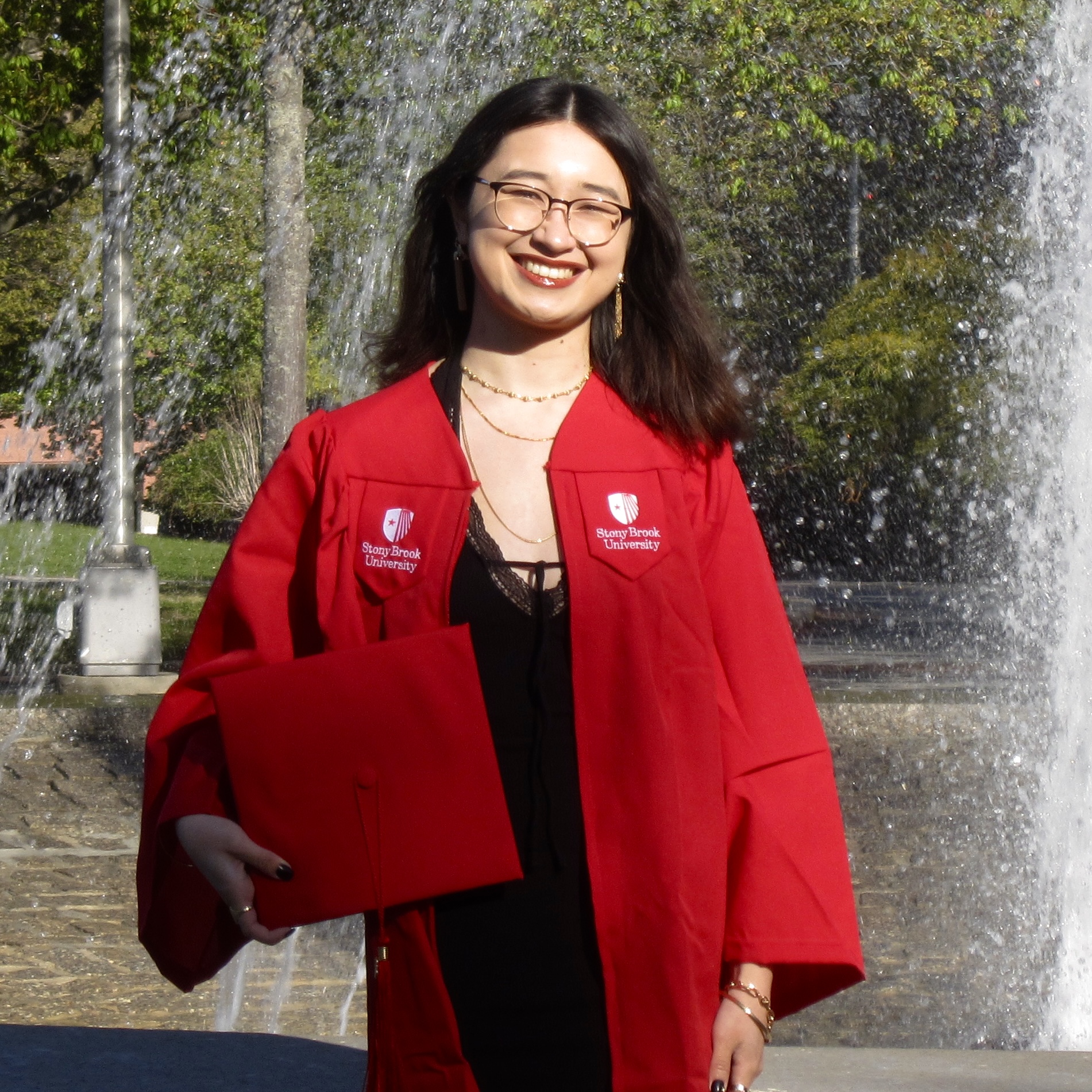 A graduate in a Stony Brook University red cap and gown stands smiling by a fountain. They hold a diploma folder, with trees and sunlight in the background. 