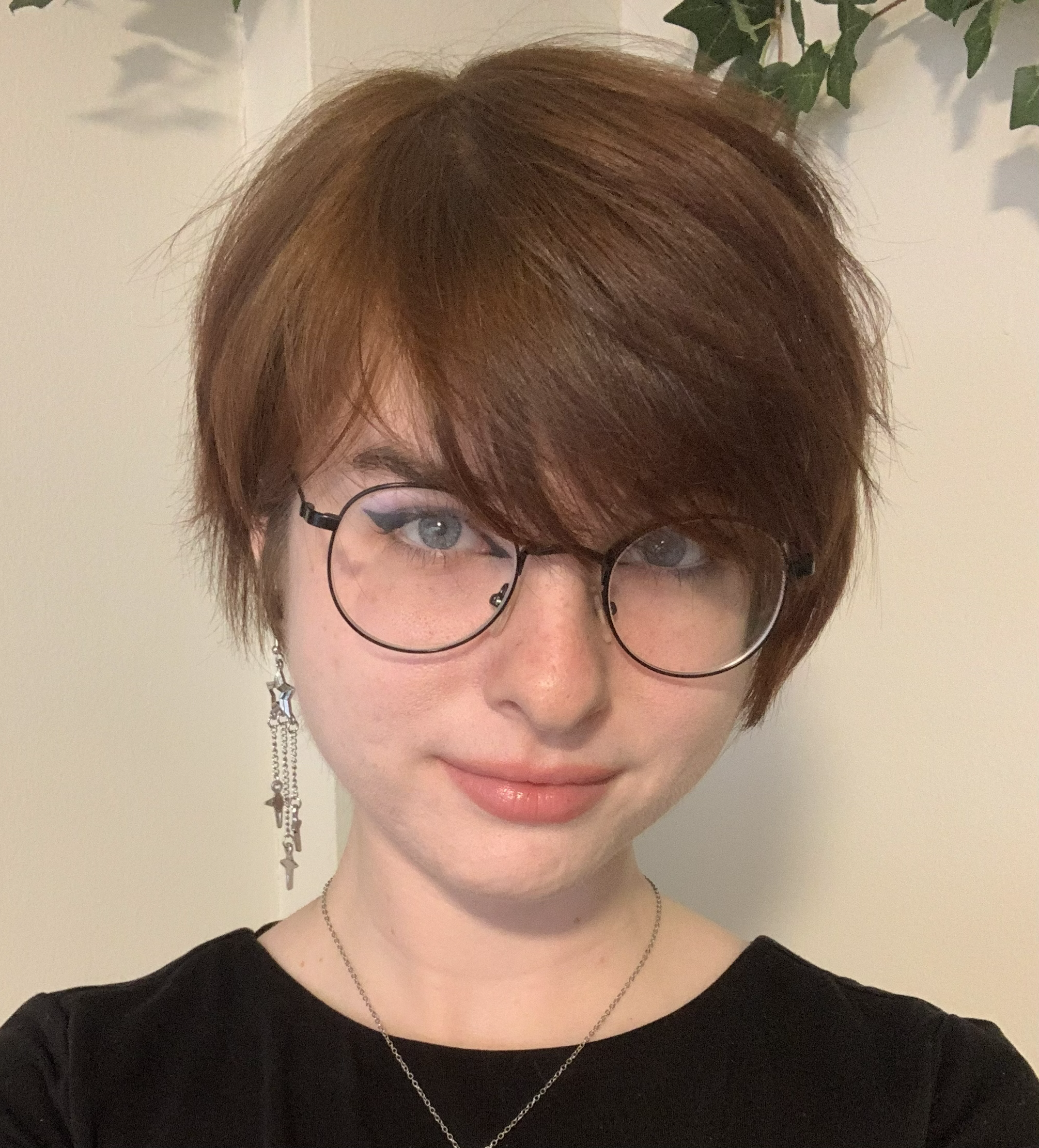 Young person with short auburn hair and glasses, wearing a black top and silver star earrings, smiles gently against a neutral background with greenery.