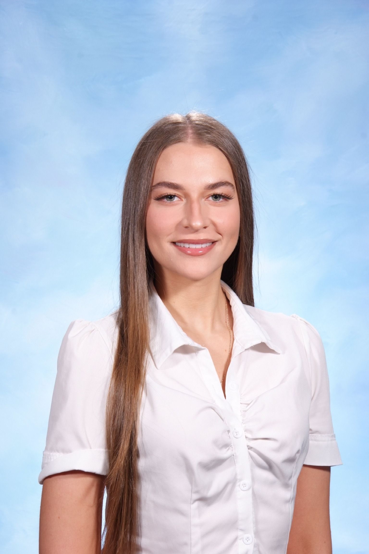 A woman with long brown hair smiles warmly against a light blue background. She wears a white blouse, creating a professional and approachable tone.