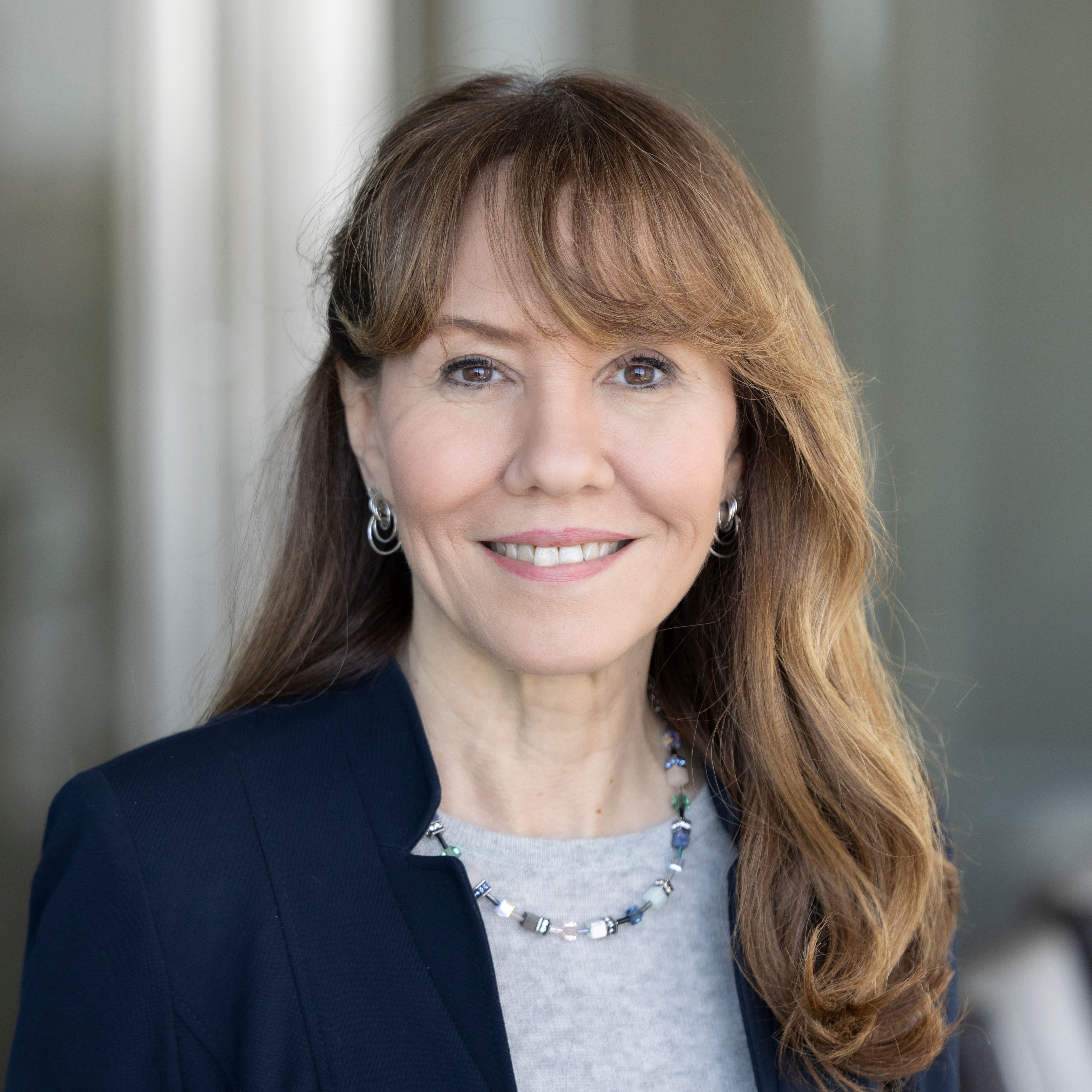 Professional portrait of a smiling individual with long brown hair, wearing a dark blazer and a silver necklace.