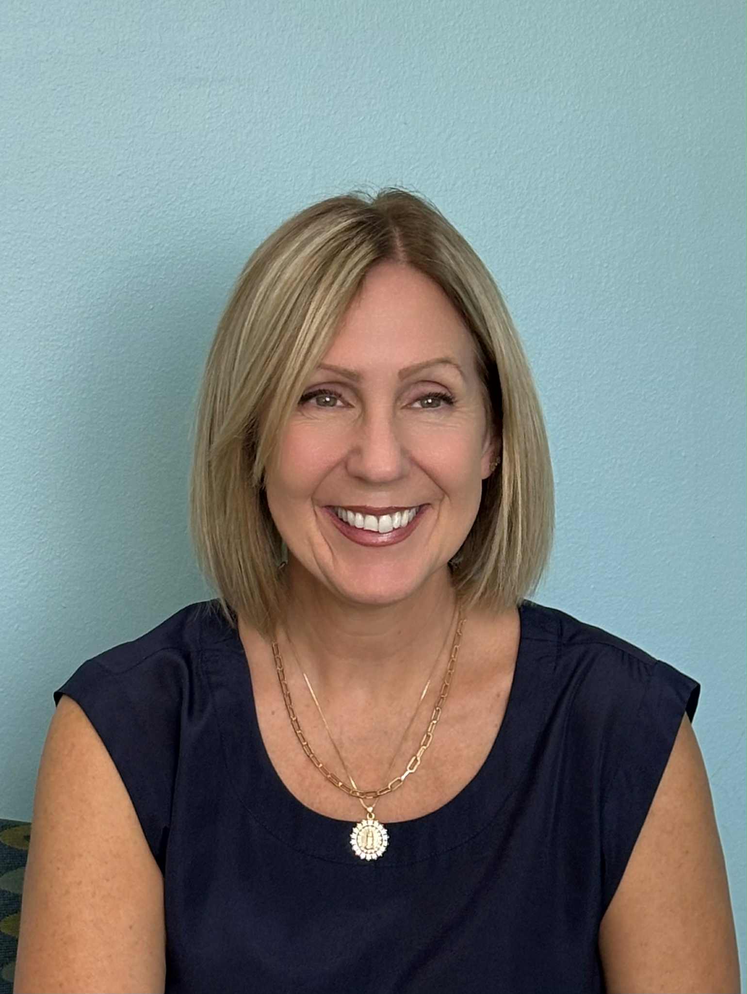 Tracey Braile A woman with blonde hair wearing a necklace sits comfortably on a couch, looking relaxed and engaged.
