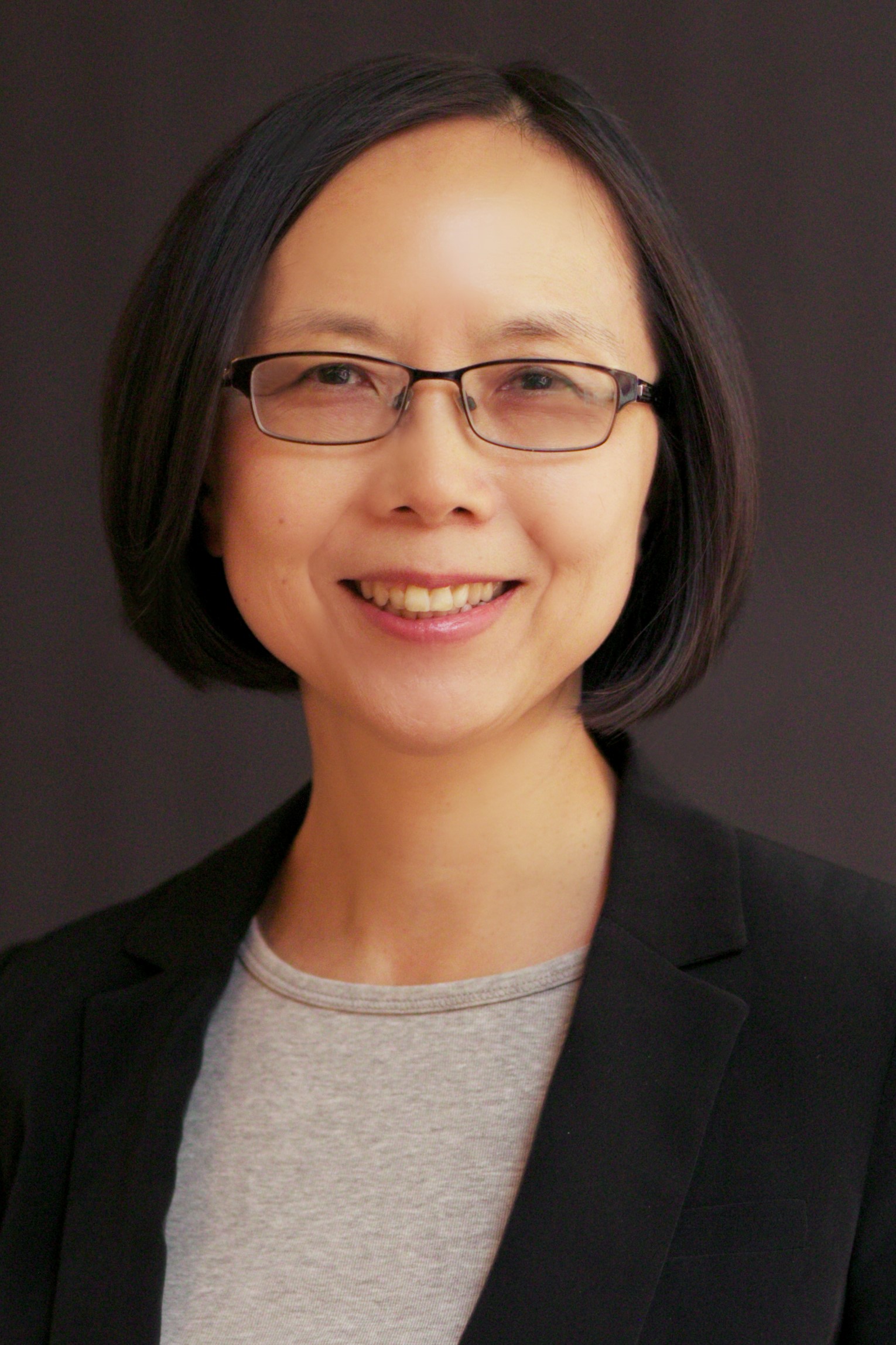 Portrait of an individual with glasses smiling, wearing a black blazer over a grey top, set against a dark background.