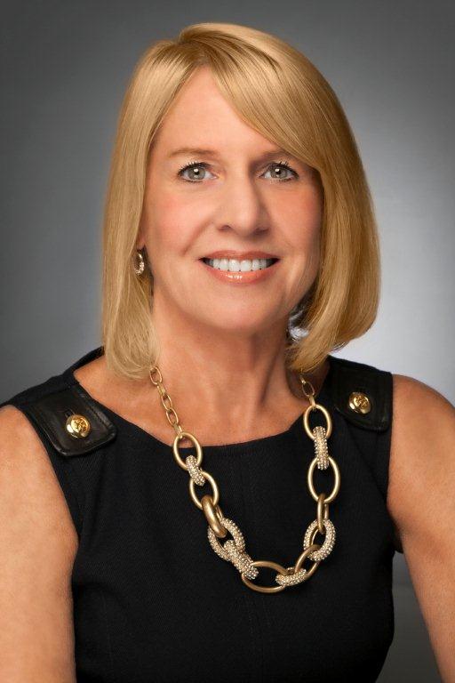 Portrait of a woman wearing a black sleeveless top and a gold chain necklace, smiling against a gray background.