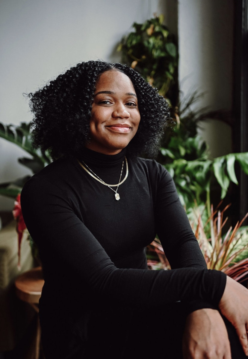 Person sitting with a smile, wearing a black turtleneck, seated indoors with plants in the background.