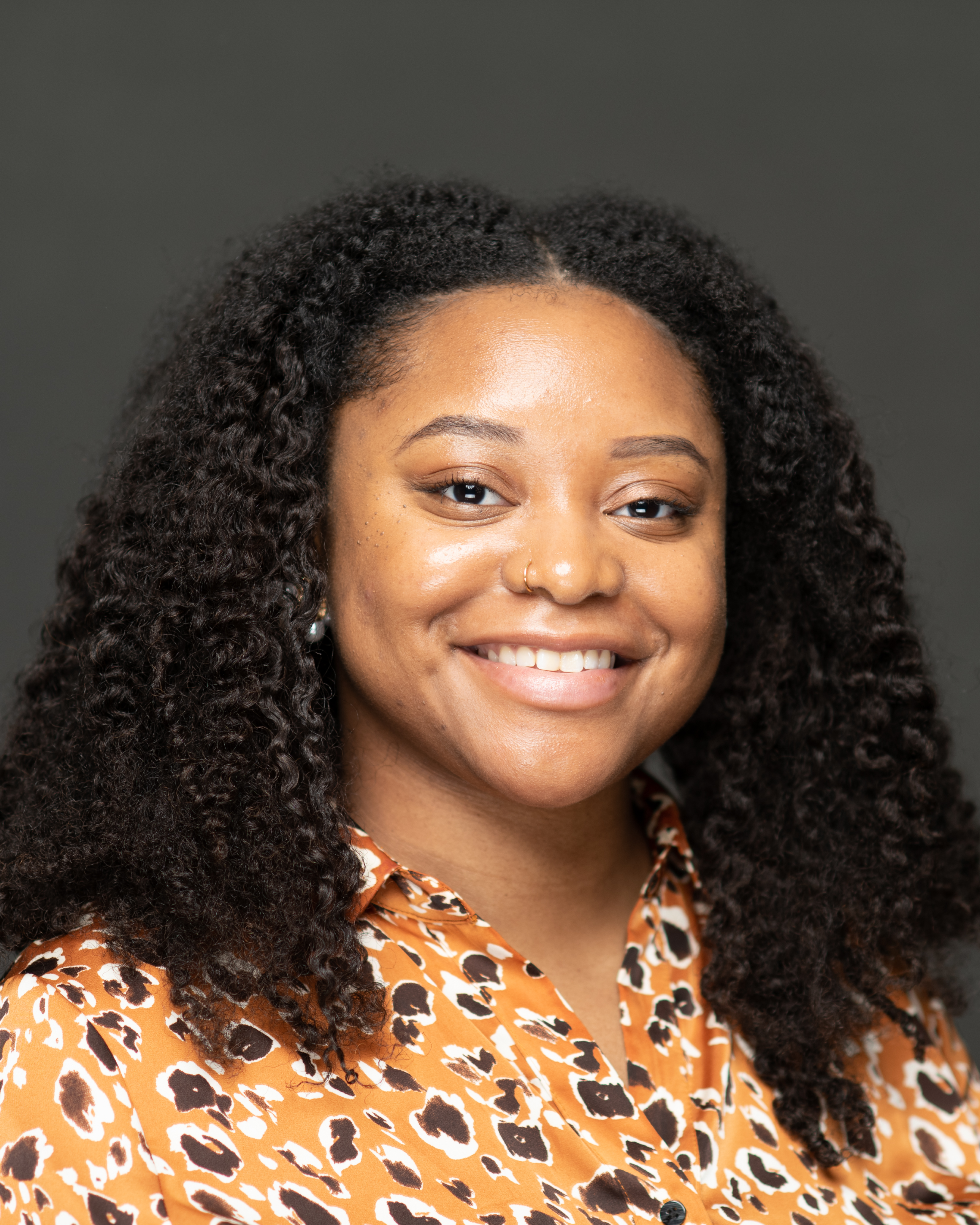 Portrait of a smiling person with curly hair, wearing a leopard print shirt, against a gray background.