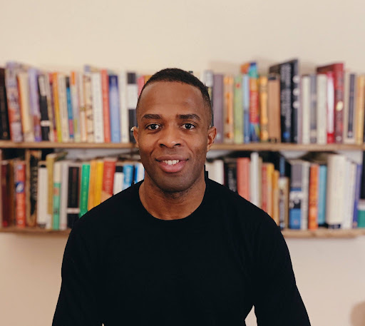 Person in black shirt smiling in front of a bookshelf filled with colorful books.