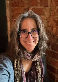 Person with glasses smiling at the camera, wearing a scarf and a patterned jacket, standing in front of a brick wall.