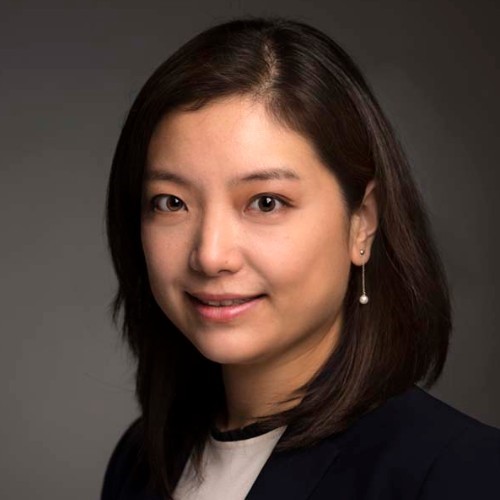 Portrait of a professional wearing a dark blazer over a light top, with subtle earrings, against a grey background.