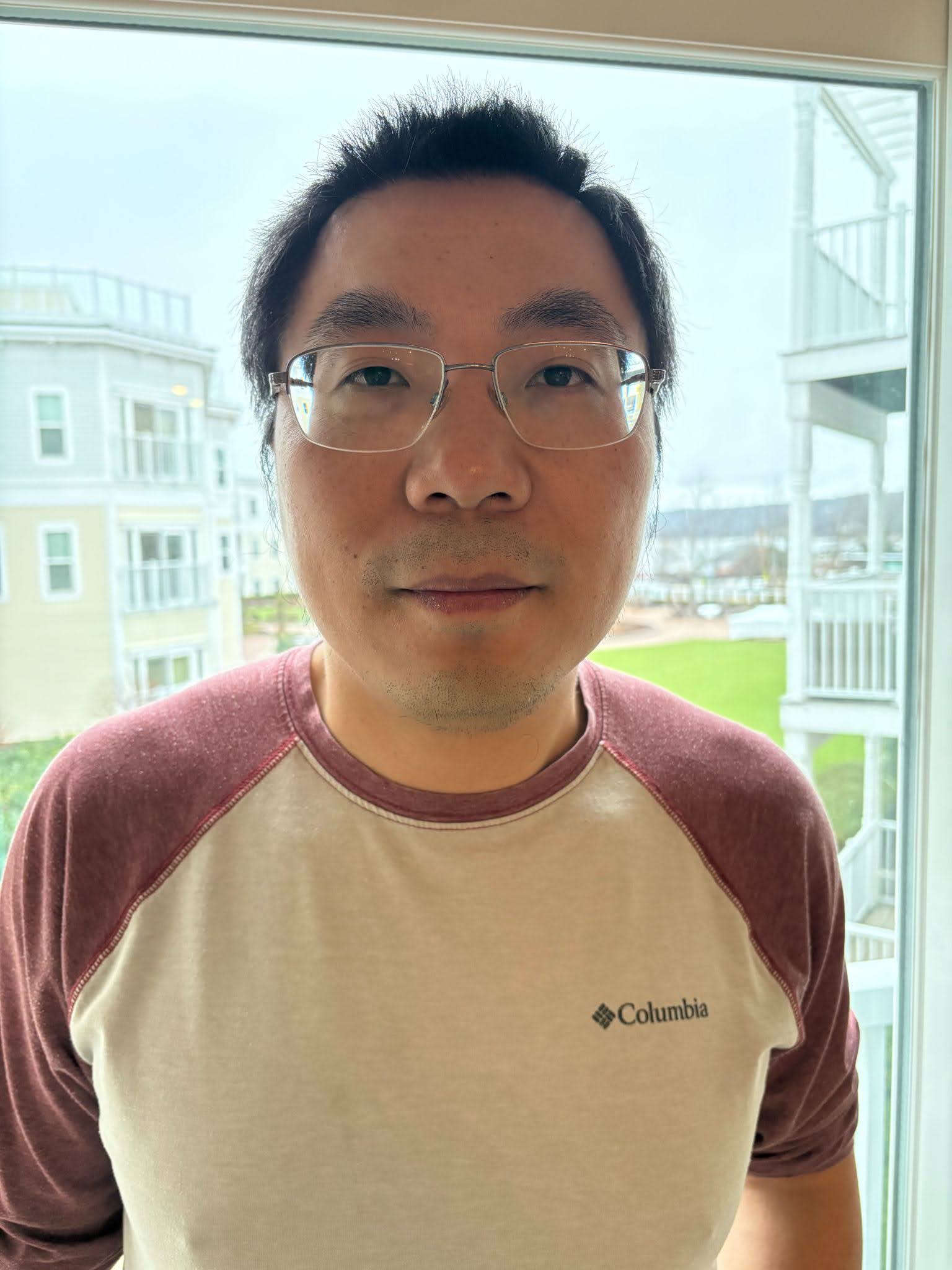 Person wearing glasses and a Columbia branded t-shirt, standing in front of a window with a view of buildings and stairs outside.
