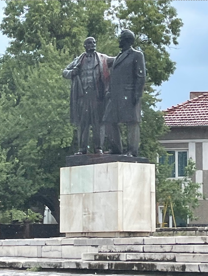 Two bronze statues of men in formal attire stand side by side on a large marble pedestal, surrounded by trees and buildings in the background.
