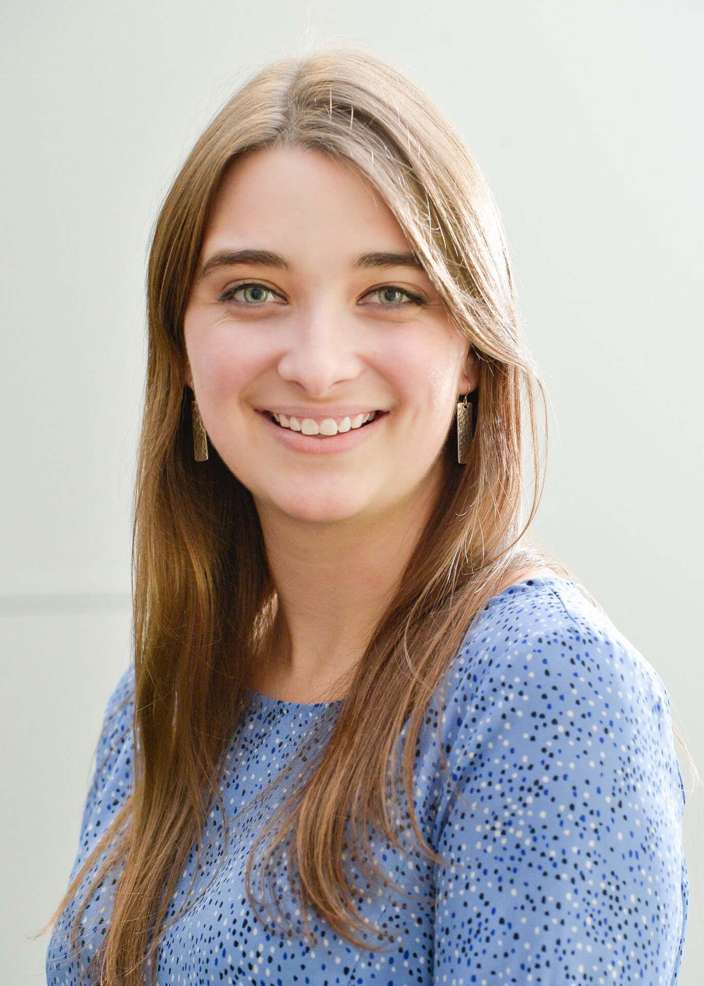 A professional headshot of a smiling young woman with long, straight light-brown hair. She is wearing a blue top with a small dark-blue speckled pattern and rectangular hanging earrings. The photo is set against a plain, neutral-colored wall.