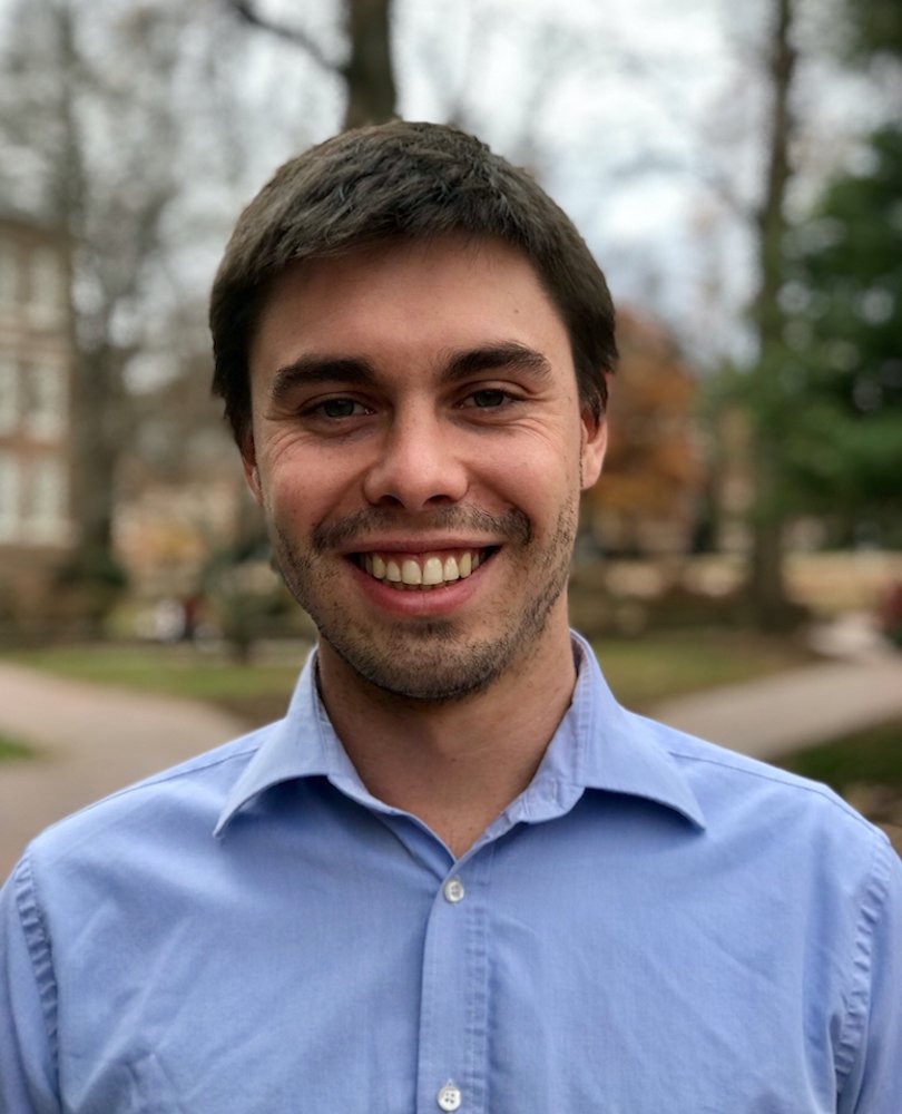Headshot of a smiling person with short dark hair wearing a light blue button down shirt, standing outdoors with trees blurred in the background.