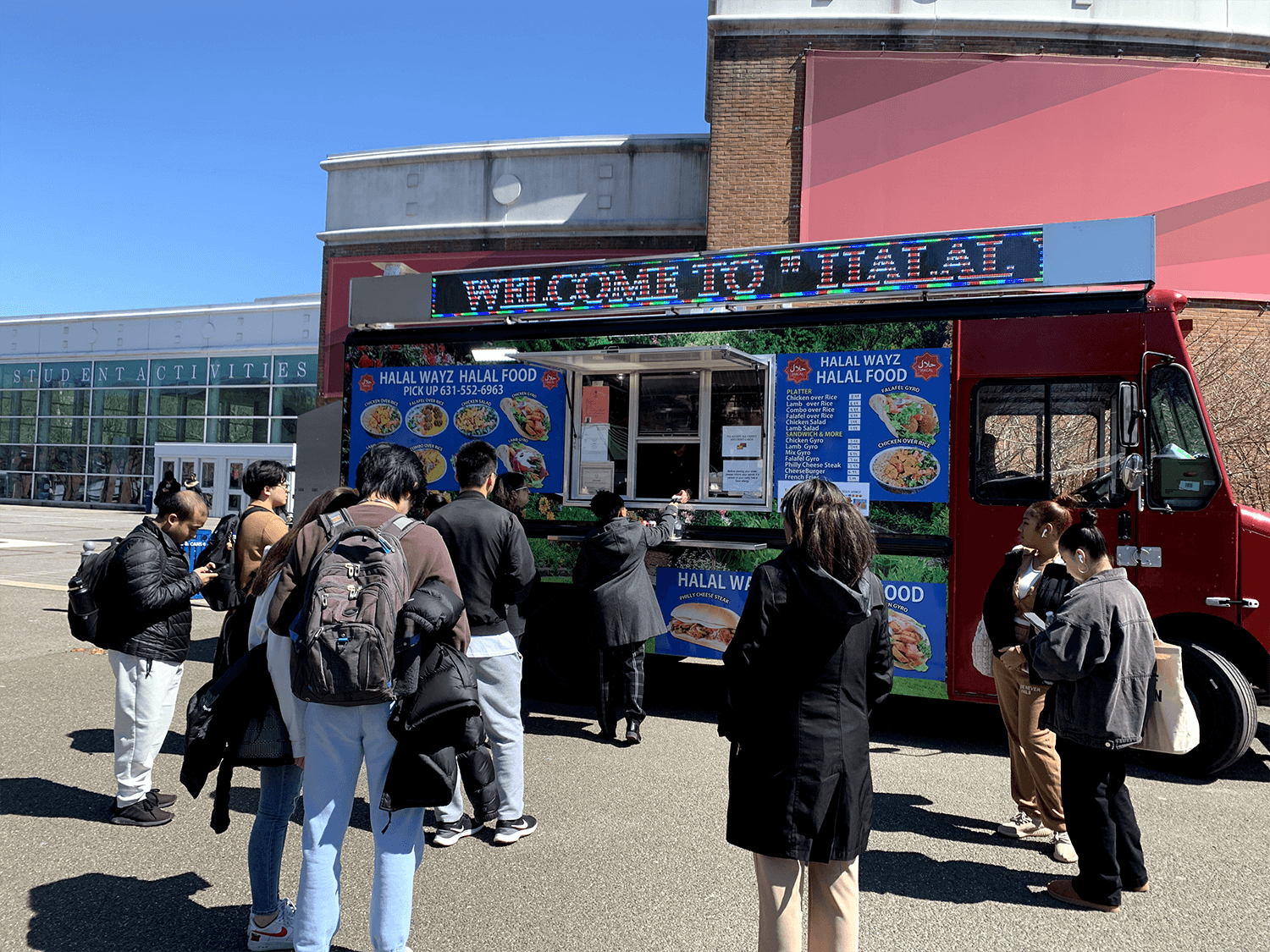 A red food truck named "HALAL WAYZ HALAL FOOD" is parked in front of a student activities building, with a line of diverse people waiting to order food on a sunny day