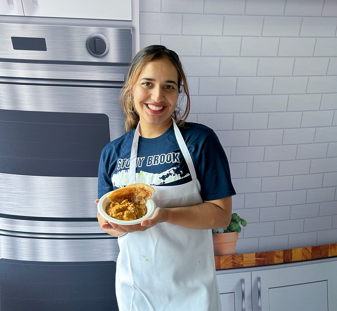 Smiling woman in a Stony Brook shirt and white apron holding a bowl of curry.