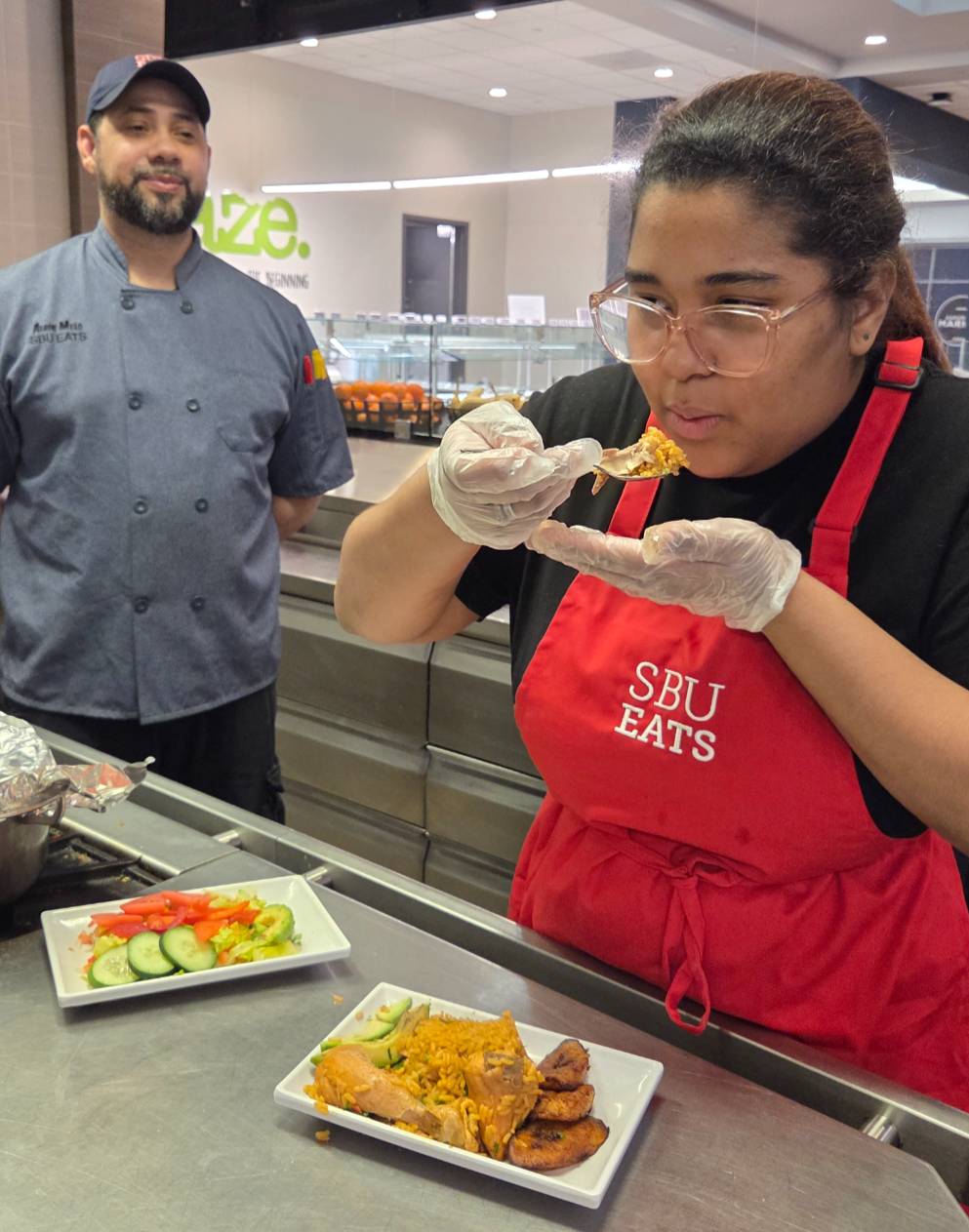 Woman in "SBU EATS" apron tasting food.