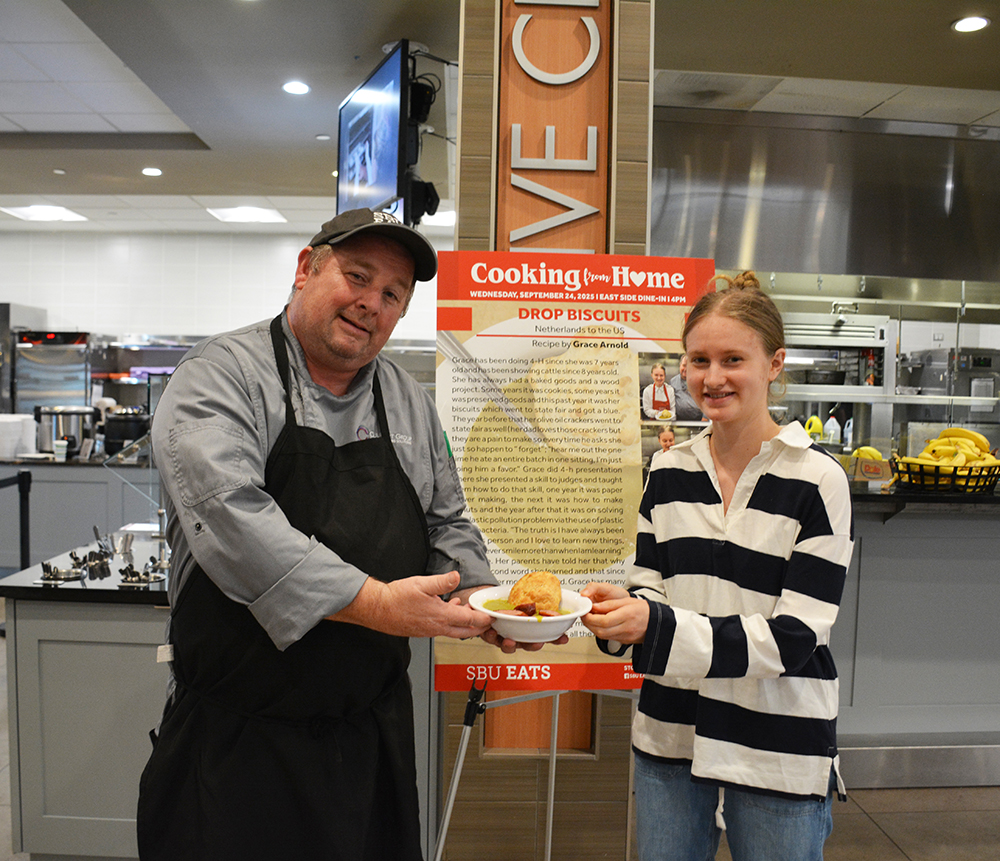 Student and chef posing with a bowl of "Drop Biscuits" by a recipe poster.