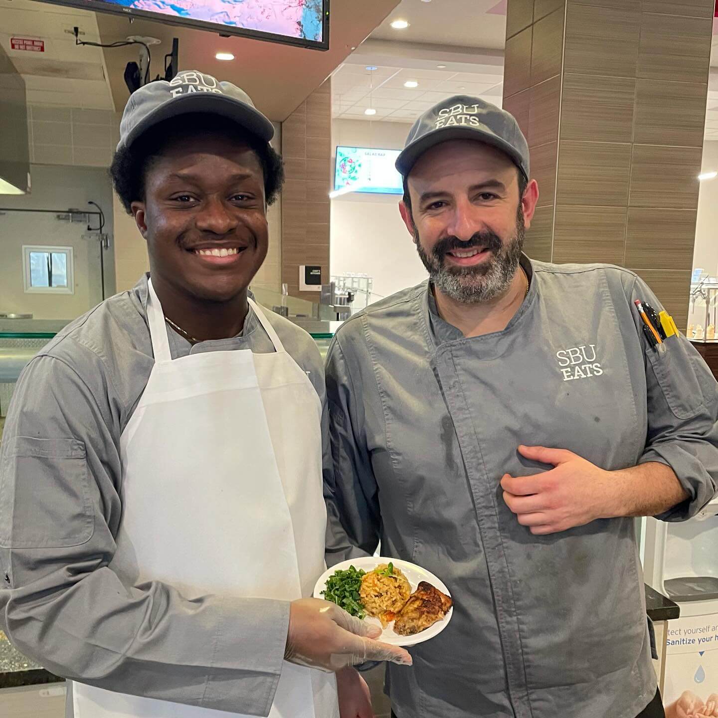 Two chefs smiling while holding a plate of chicken, rice, and greens.